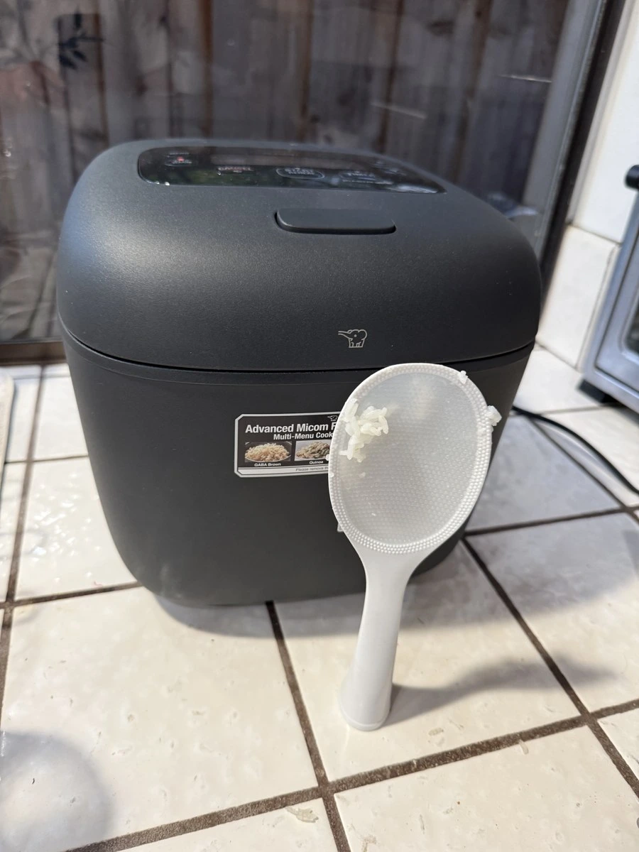 My Zojirushi self-standing white embossed rice paddle (shamoji) standing upright on the counter in front of the NL-EAC18BA with a few rice grains sticking to the paddle